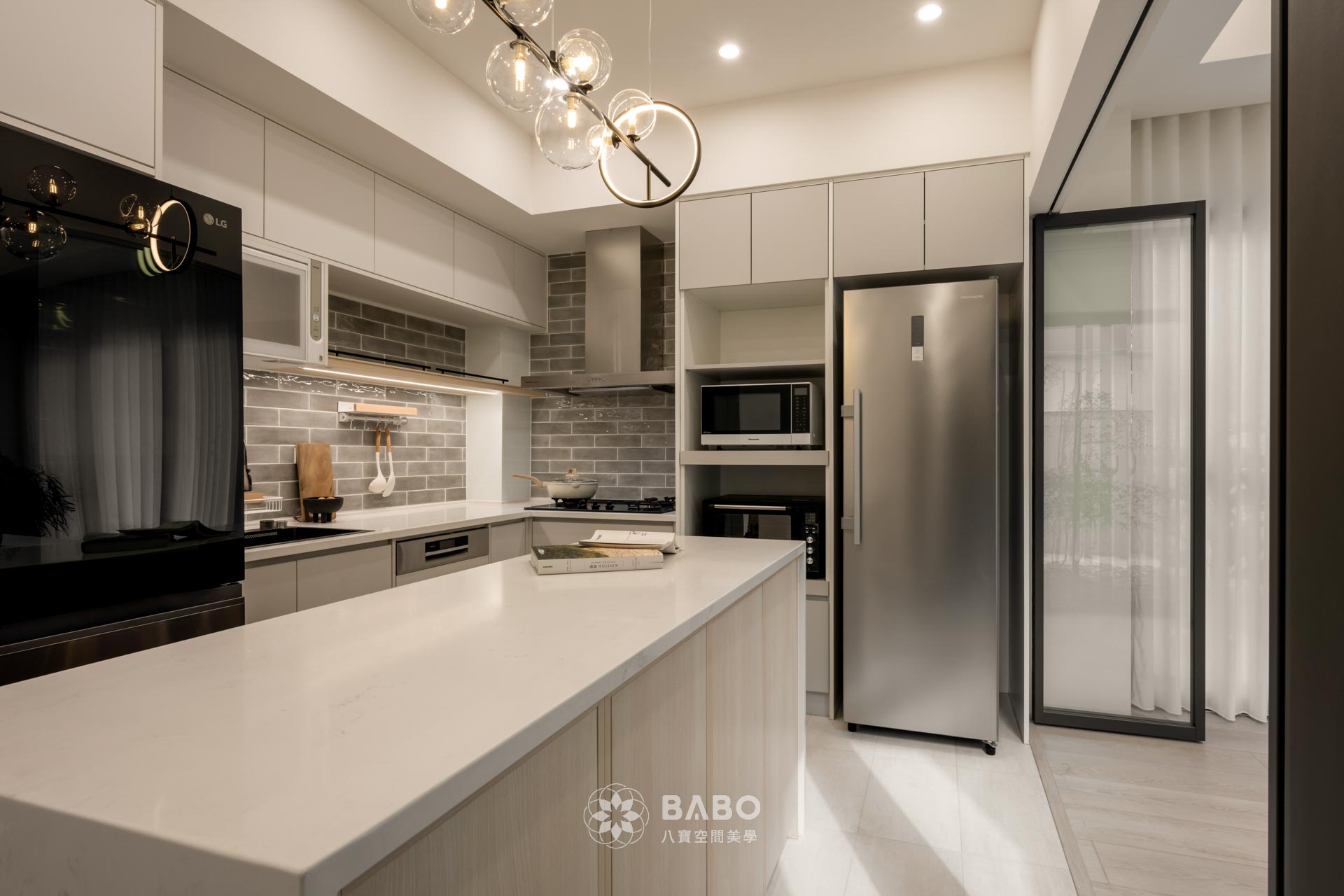 ▲廚房空間也是整體設計中的一大亮點,採用灰色亮面壁磚、白色石英石檯面與深灰色一體式水槽,營造出純淨的烹飪氛圍。中央大中島不僅具備備餐與收納功能,也串連起生活交流與視覺焦點,成為日常生活中不可或缺的靈魂角落。